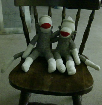 Two sock monkeys on a chair, neither yet having eyes.