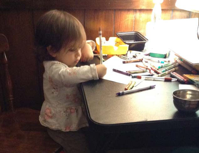 Toddler seated at a card table using pencils and crayons.